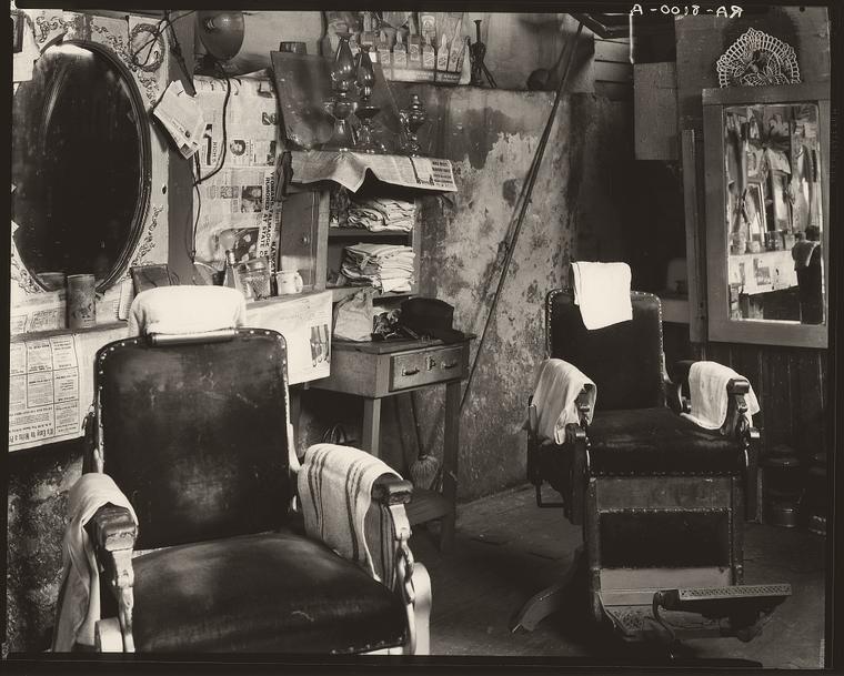 Archival photograph: the interior of an Atlanta barber shop in 1936, two empty barber chairs with folded towels over the arms, an oval mirror and a swept wooden floor.