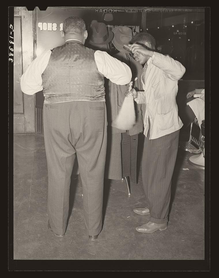 Archival photograph: a barber brushing hair off a customer's suit at the door of a Southside Chicago barber shop, 1941, hats on the rack behind them.