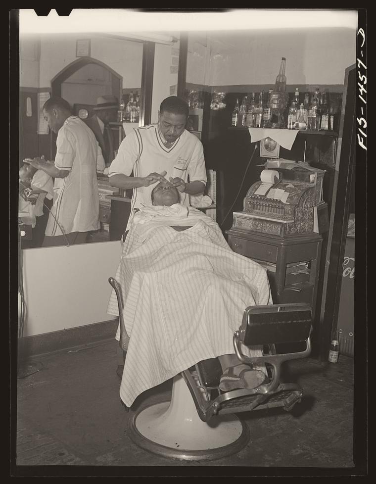 Archival photograph: barber Oscar J. Freeman in a white smock working on a reclined customer in the Metropolitan Barber Shop, Chicago, 1942, an ornate register on the counter beside him.