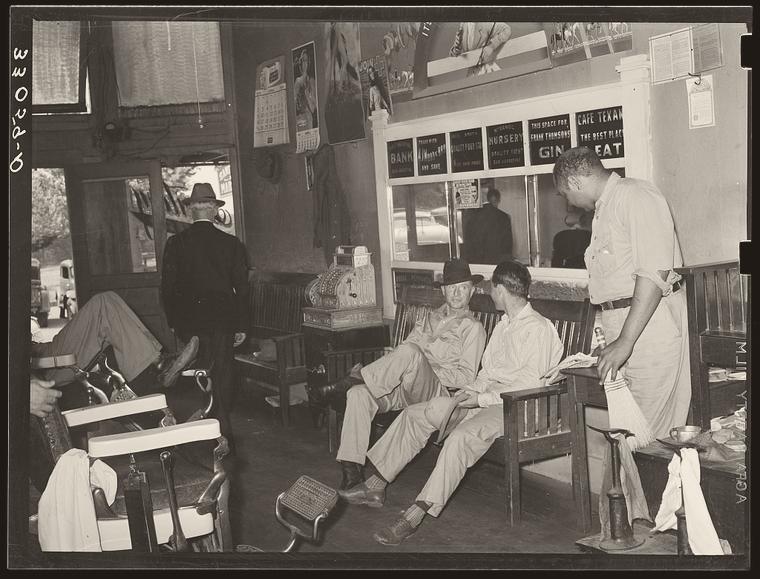 Archival photograph: men sitting and waiting on a bench inside a San Augustine, Texas barber shop on a Saturday afternoon, 1939, one barber working at the chair.
