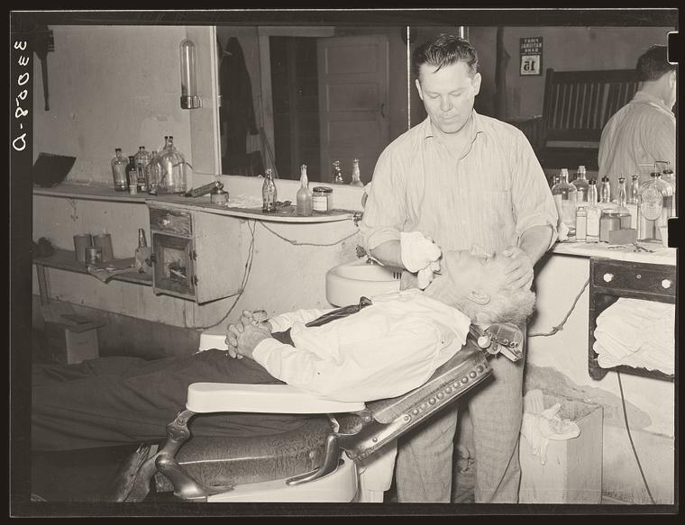 Archival photograph: a barber leaning in close, applying the finishing touches to a customer's shave in San Augustine, Texas, 1939.