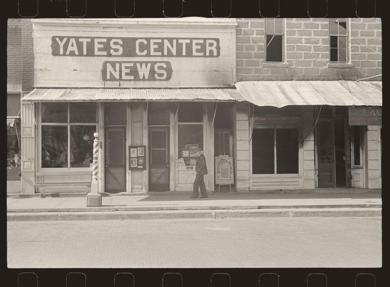 Archival photograph: a small-town storefront row in Yates Center, Kansas, 1938, holding a barber shop with a striped pole, a newspaper office and a photographer's studio.